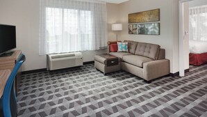 A hotel room with a sofa, a TV, a lamp, and a large window with sheer curtains. - TownePlace Suites by Marriott Titusville Kennedy Space Center (Titusville)