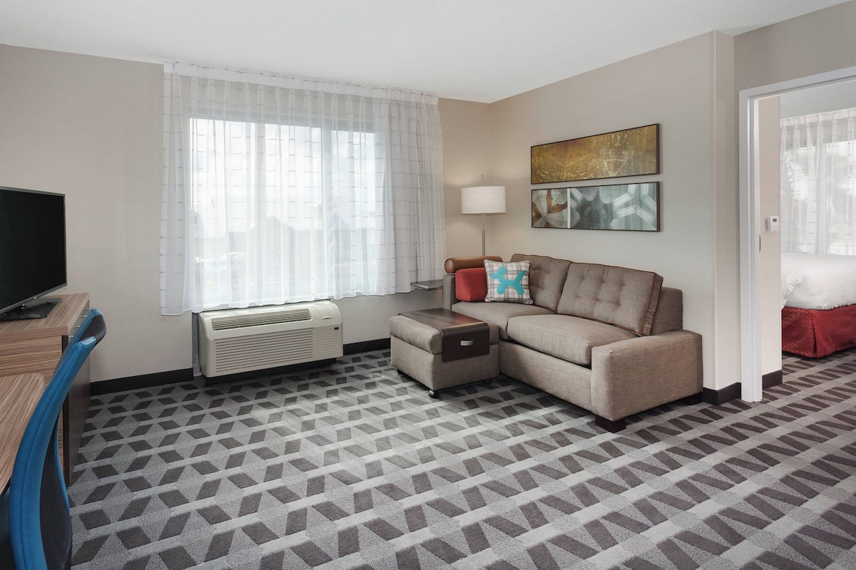 A hotel room with a sofa, a TV, a lamp, and a large window with sheer curtains.