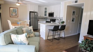 Eine moderne Küche mit weißen Schränken, einem Edelstahlkühlschrank und einer Mikrowelle. Es gibt eine Essecke mit Tisch und Stühlen, sowie ein Wohnzimmer mit einer Couch und einem Couchtisch.