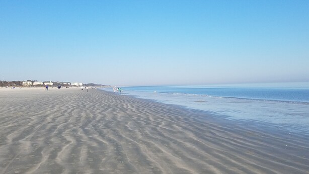 Ein weiter Sandstrand mit sanften Wellen, blauem Himmel und Gebäuden in der Ferne.