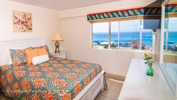Un dormitorio con cama, mesita de noche, lámpara, ventana con vista al mar y una pequeña mesa con un florero.