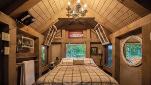 Ein gemütliches Schlafzimmer im Dachgeschoss mit einem Bett, einer Kristallleuchtre, einem Fenster mit Blick auf Bäume und einem Spiegel an der Wand.