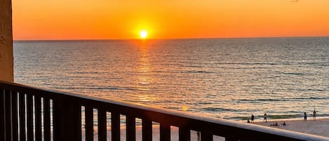 A balcony with a view of the sunset over the ocean, a table, and a railing.