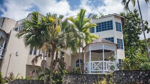 A multi-story building with a white exterior, a covered outdoor area with a lattice roof, and surrounding tropical vegetation. - Spectaculer View at Columbus Heights (Ocho Rios)