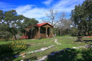 A small wooden cabin with a red roof, surrounded by a grassy area with a stone pathway leading to it. - 1 Room Cabin getaway, quiet country views, wildlife, pet friendly (Kerrville)