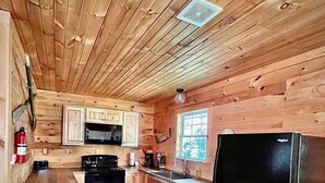 Une cuisine rustique, située dans une cabane en bois, avec un réfrigérateur noir, des placards en bois, un four à micro-ondes et une fenêtre.