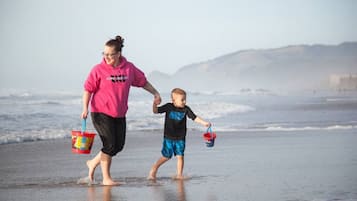 A person and a child walking on a beach holding hands, with a clear view of the ocean and distant hills.