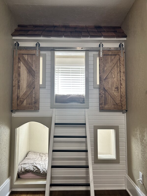 A loft bedroom with a loft bed, a window with blinds, and a ladder.