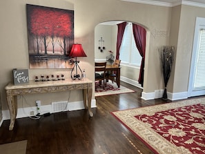 A living room with a wooden console table, a red lamp, a painting of a red tree, a dining area with a table and chairs, and a red patterned rug.