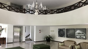A spacious lobby with a chandelier, a staircase, and a seating area with two green ottomans and a wicker sofa. - Apartamento Porto Praia do Forte (Mata de São João)