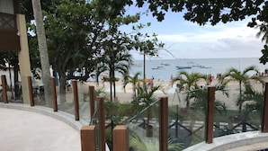A pool area with a glass barrier, palm trees, and a view of the beach with boats. - Apartamento Porto Praia do Forte (Mata de São João)