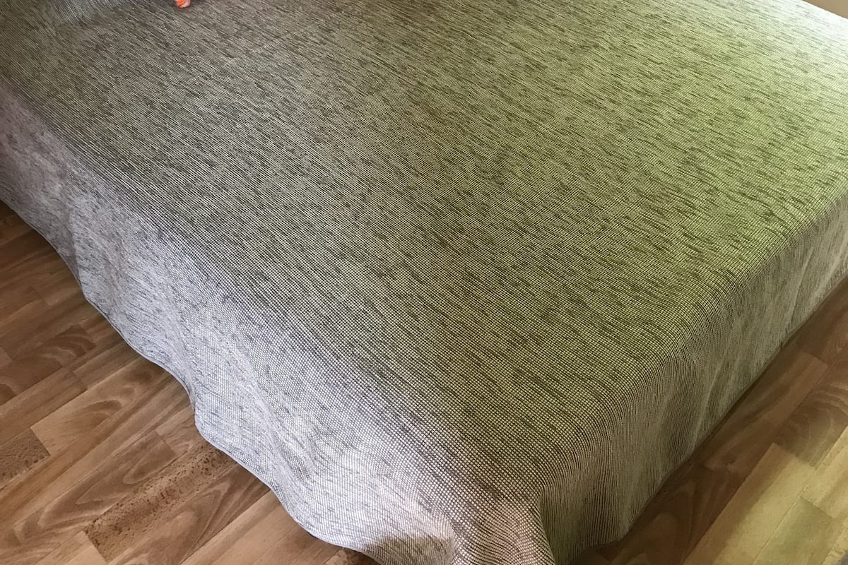 A single bed with a grey and green textured bedspread, an orange patterned pillow, and a wooden floor.