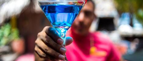 A person holding a blue cocktail glass with a garnish, in an outdoor setting with a thatched roof structure in the background.