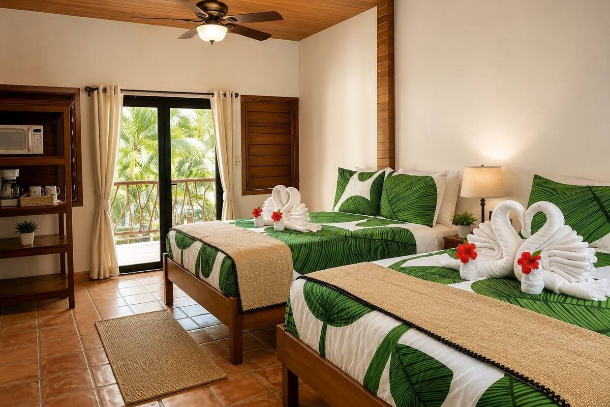 A hotel room with two beds, a ceiling fan, a balcony with curtains, a wooden shelf with a microwave, and a view of palm trees.