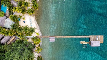 An aerial view of a beach with a wooden pier connecting to a bungalow with a balcony.
