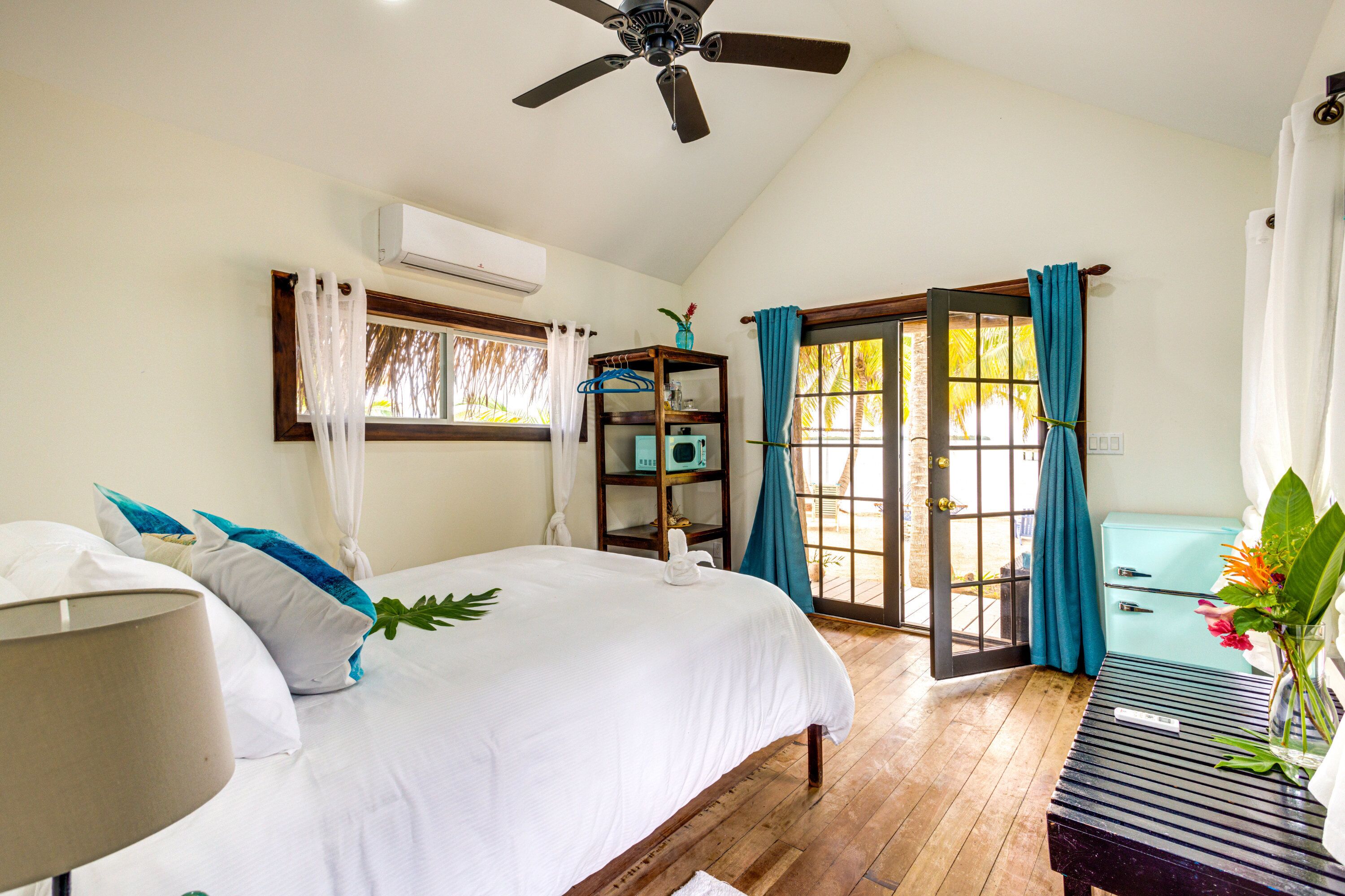 A bedroom with a bed, a bedside table, a ceiling fan, a window with curtains, and a view of the outdoors.