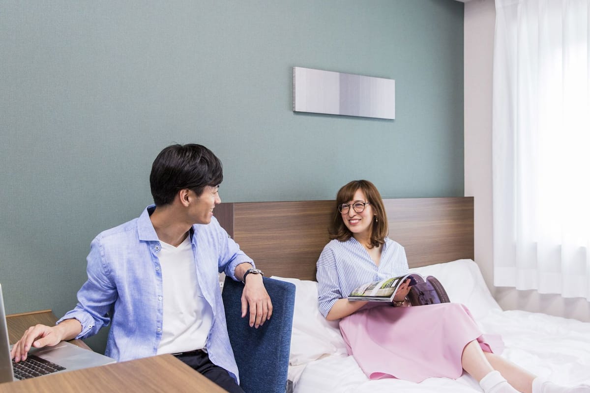 A hotel room with a bed, a bedside table, a laptop, and a person reading a book.