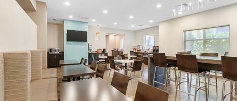 Un espace repas moderne doté de plusieurs tables et chaises, d’un coin cuisine et d’une grande fenêtre laissant entrer la lumière naturelle.