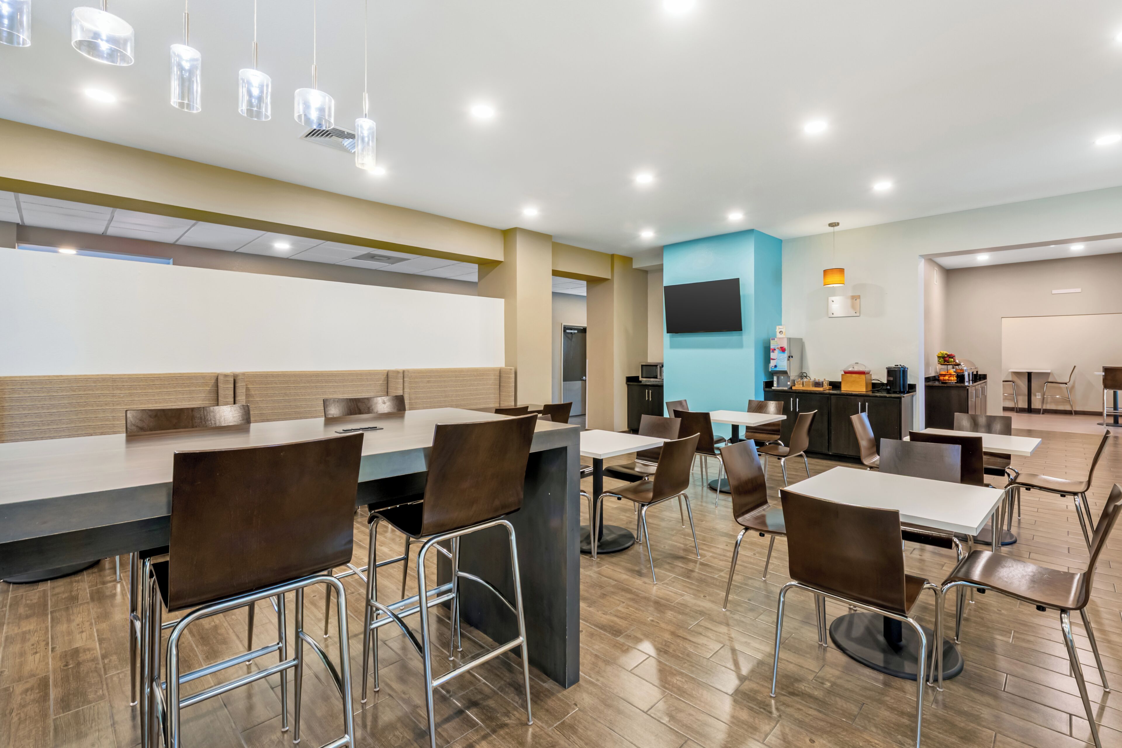 Un espace repas moderne avec des tables et des chaises hautes, un téléviseur à écran plat fixé au mur et un coin cuisine équipé d’appareils électroménagers et d’un présentoir à fruits.
