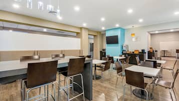 Un espace repas moderne avec des tables et des chaises hautes, un téléviseur à écran plat fixé au mur et un coin cuisine équipé d’appareils électroménagers et d’un présentoir à fruits.