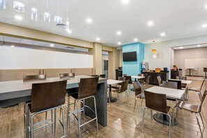 A modern dining area with high tables and chairs, a flat-screen TV mounted on the wall, and a kitchen area with appliances and a fruit display. - Caretta Landing Sarasota, an Ascend Collection Hotel (Sarasota)
