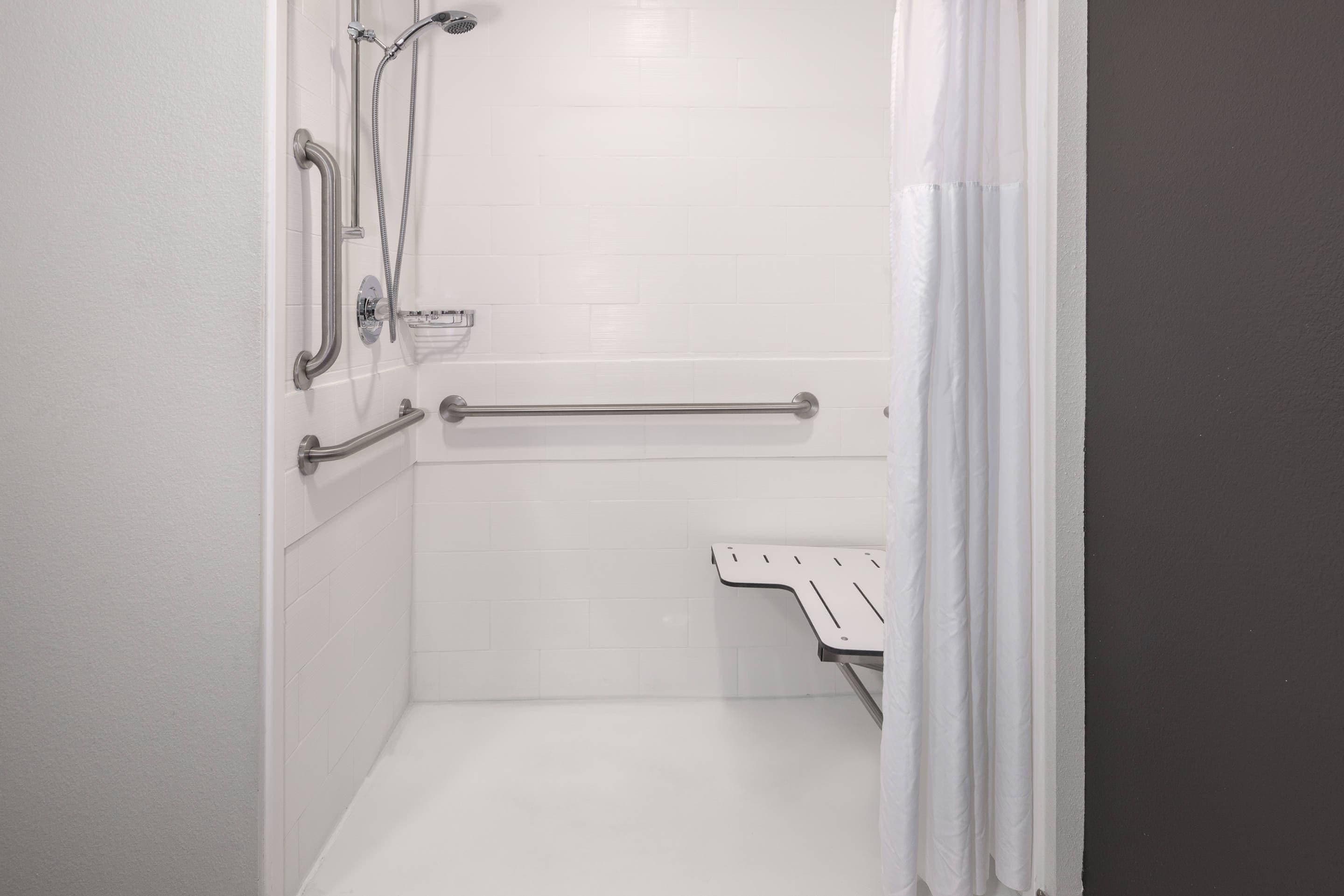 A bathroom with white tiles, a shower with a handheld showerhead, a grab bar, and a white shower seat.