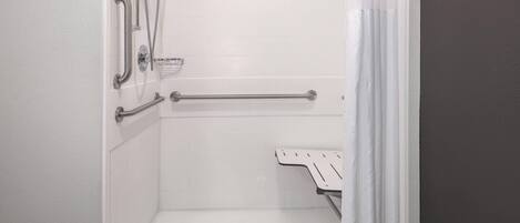 Une salle de bain carrelée de carreaux blancs, une douche équipée d’un pommeau de douche à main, d’une barre d’appui et d’un siège de douche blanc.