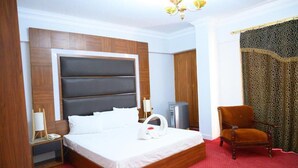 A hotel room with a large bed, a wooden headboard, a red carpet, a small desk with a lamp, a chair, and a wardrobe.