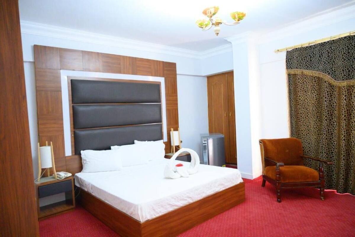 Una habitación de hotel con una cama grande, cabecera de madera, alfombra roja, un escritorio pequeño con lámpara, una silla y un armario.