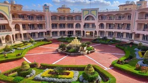 A large, multi-story hotel with a central courtyard featuring a fountain, surrounded by well-manicured gardens and a red-tiled floor.