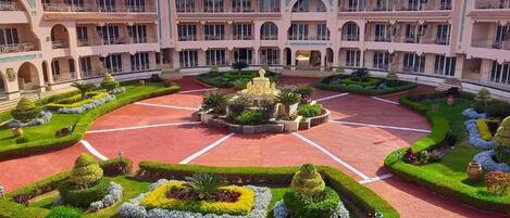 A large, multi-story hotel with a central courtyard featuring a fountain, surrounded by well-manicured gardens and a red-tiled floor.