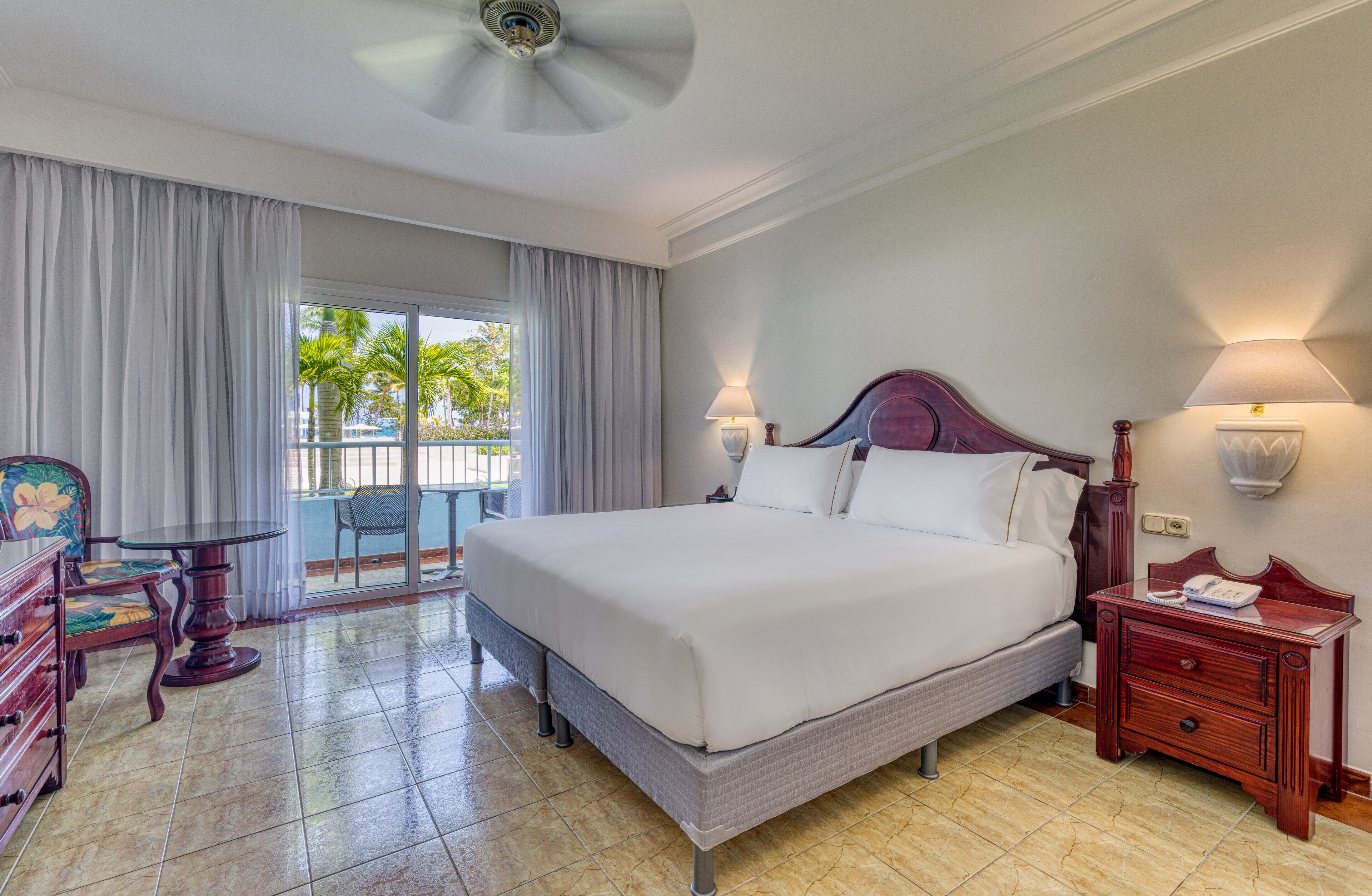 Ein Hotelzimmer mit einem großen Bett, einem kleinen Tisch, einem Stuhl und einem Balkon mit Blick auf Palmen.