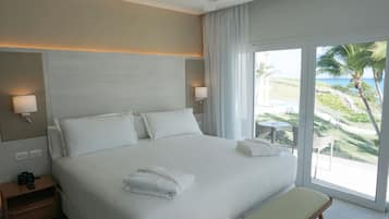 Habitación de hotel con una cama grande, lámparas de noche, mesita de noche, un banco y vistas al océano y palmeras.