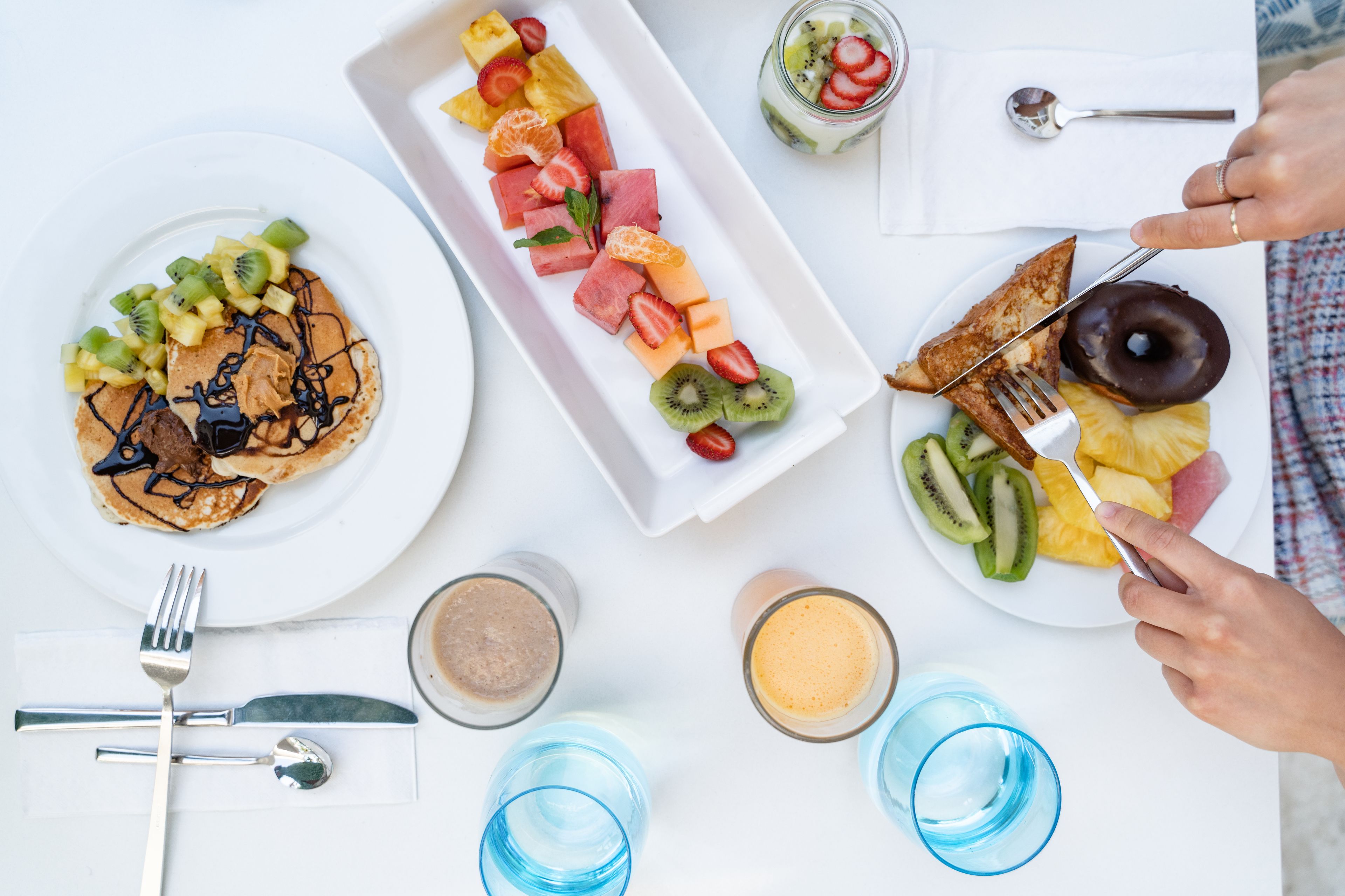 Ein Frühstückstisch mit Pfannkuchen, Obst und einem Donut auf Tellern, zusammen mit Tassen und Besteck.