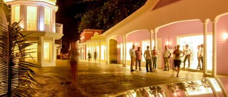 A well-lit outdoor area with a covered walkway, palm trees, and a group of people standing and conversing.