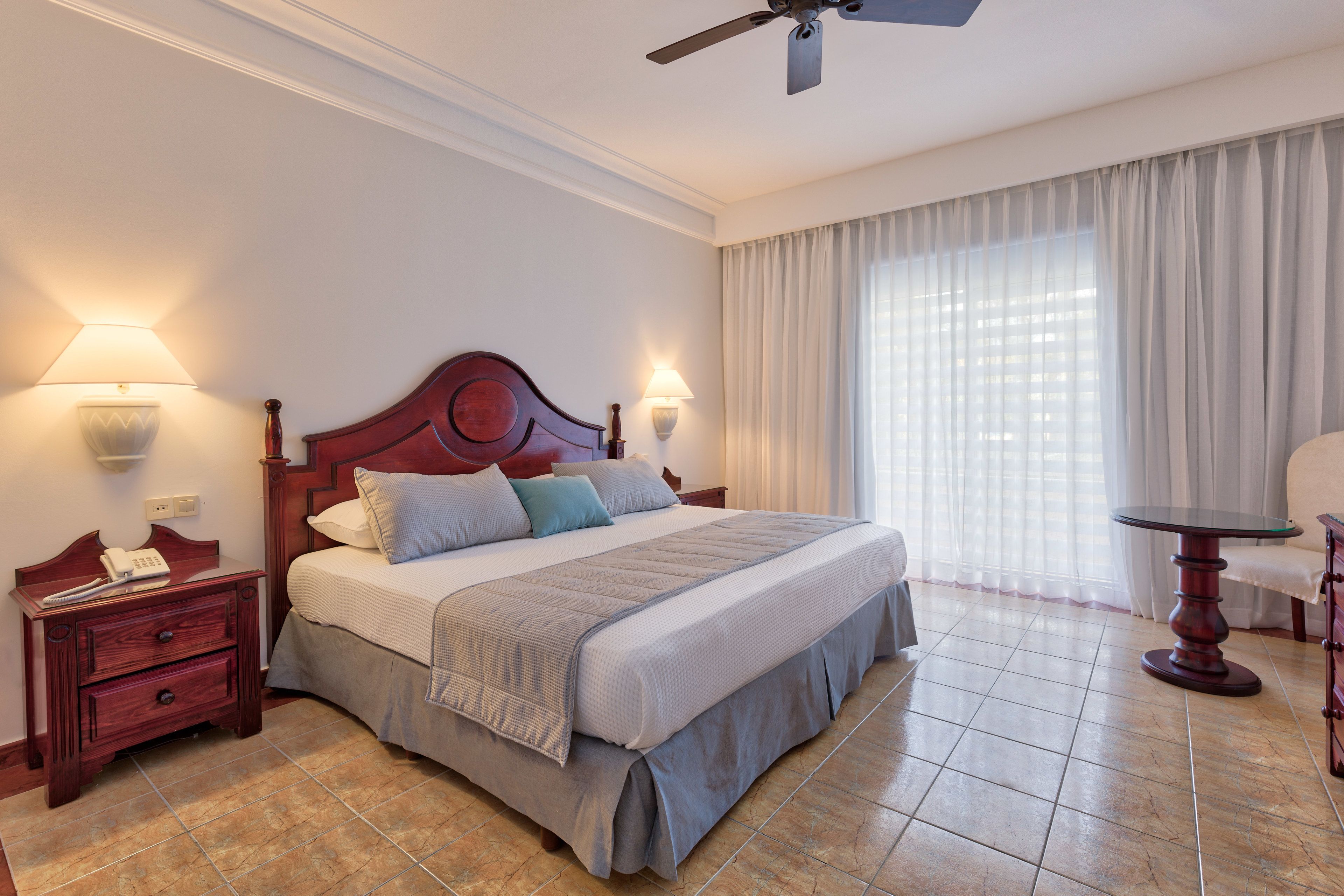 A hotel room with a large bed, two bedside lamps, a nightstand with a phone, a ceiling fan, and a window with curtains.