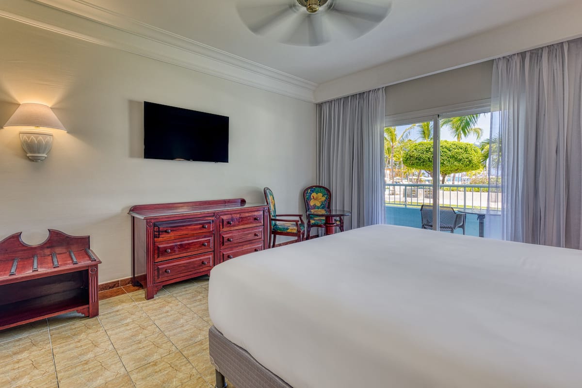 Ein Hotelzimmer mit einem großen Bett, einer Holzbank, einem Schminktisch, einem Fernseher und Blick auf einen Pool und Palmen durch das Fenster.