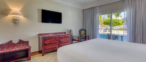 Une chambre d’hôtel avec un grand lit, un banc en bois, une commode, une télévision et une vue sur une piscine et des palmiers, visible par la fenêtre.