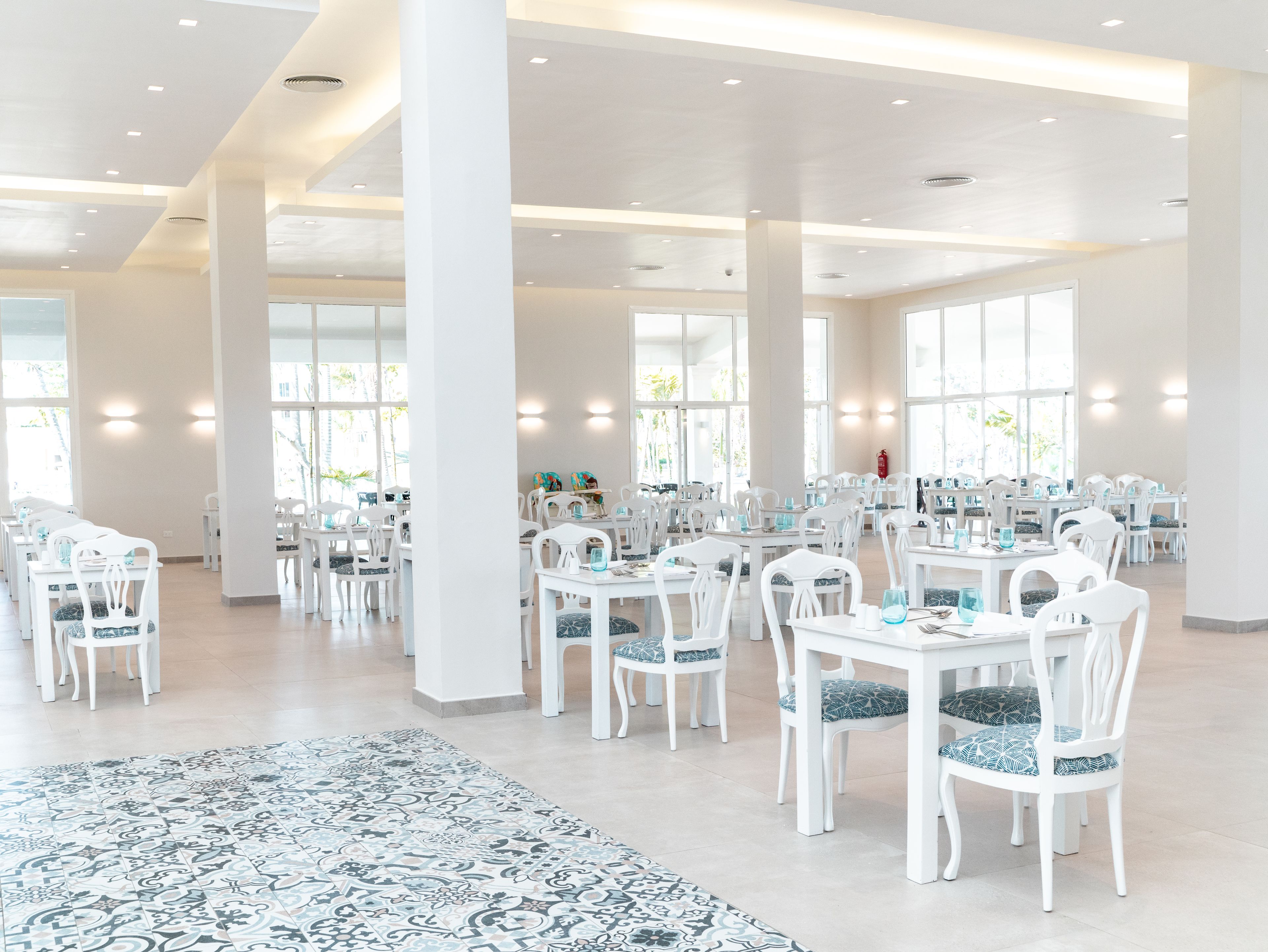 Une salle à manger spacieuse et bien éclairée, avec des tables et des chaises blanches, de grandes fenêtres et un tapis à motifs.
