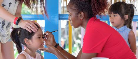A person applying makeup to another person at a table with various items on it.