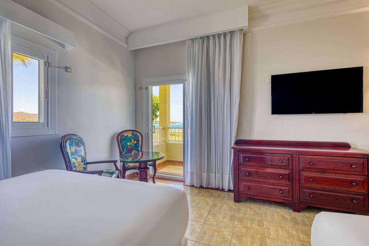 Ein Hotelzimmer mit Bett, Wandfernseher, Holzkommode, Stuhl und einem Balkon mit Vorhängen.