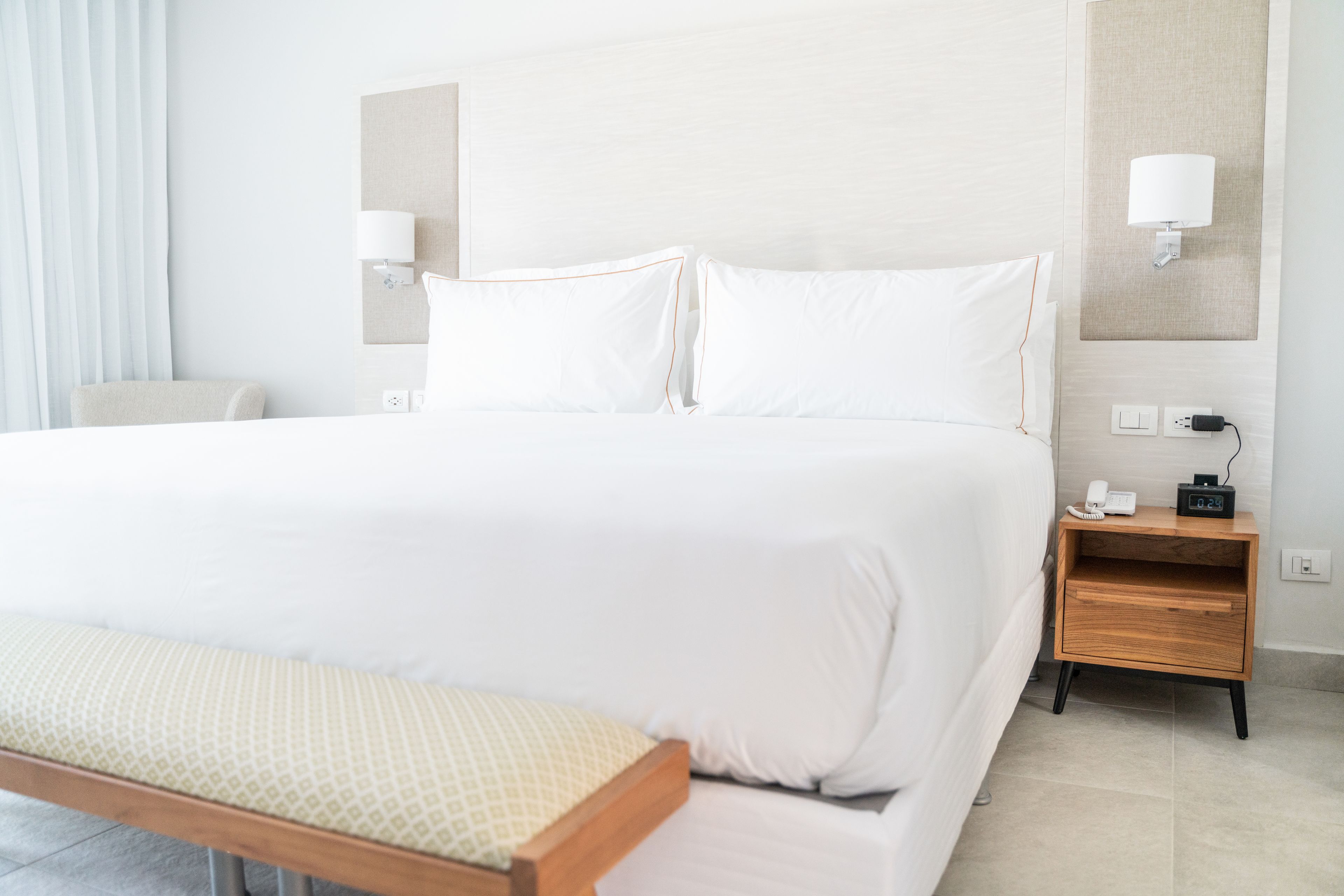 Una habitación de hotel con una cama grande, mesitas de noche, un banco y una lámpara fijada a la pared.