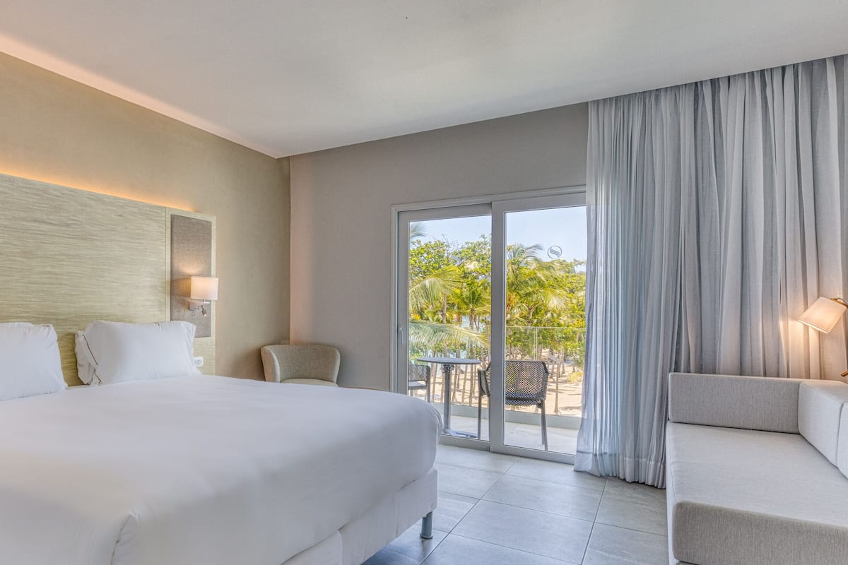 Ein Hotelzimmer mit einem großen Bett, einem Schreibtisch mit Telefon, einem Stuhl und einem Balkon mit Blick auf Palmen.