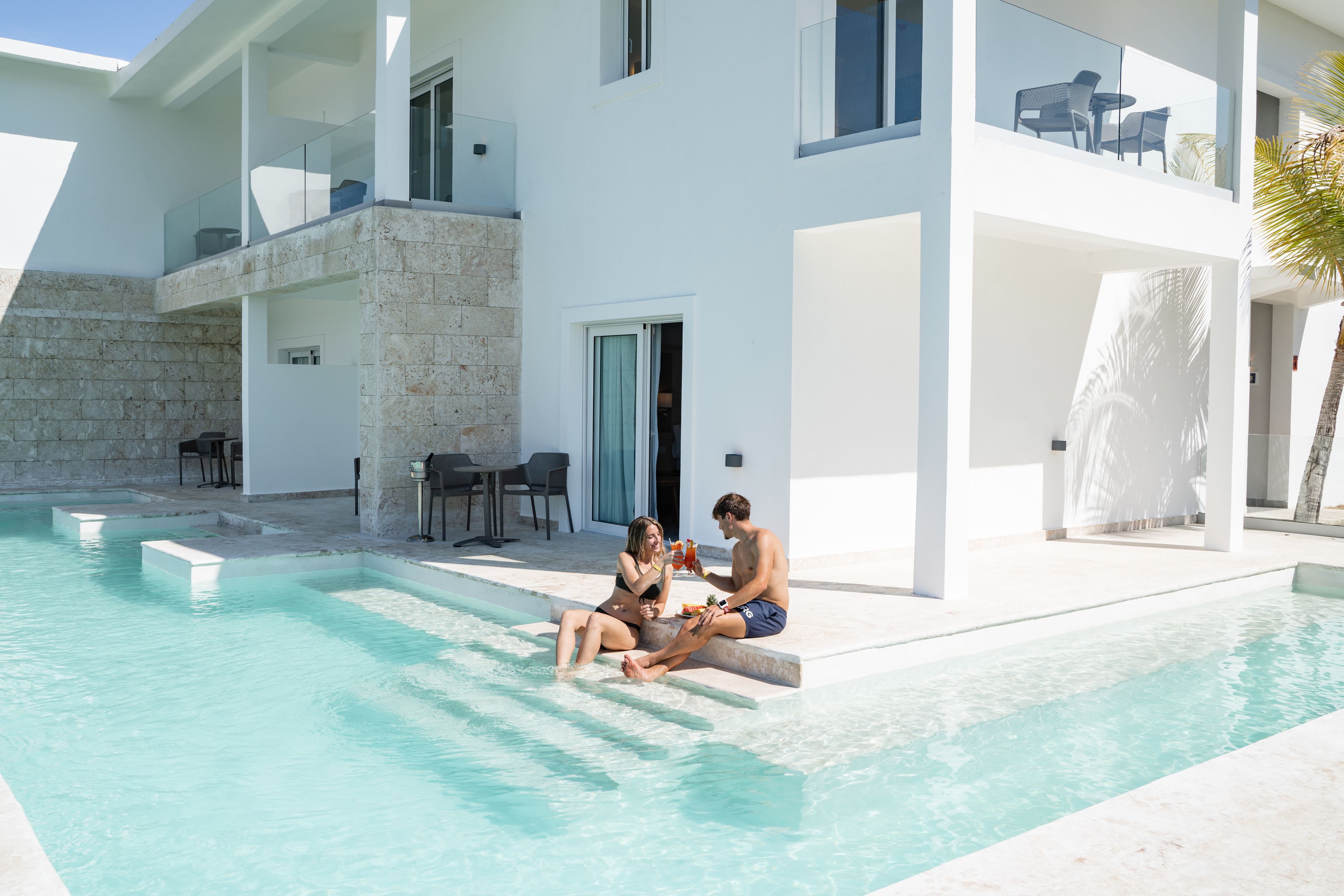 A modern pool area with clear water, a stone wall, and a two-story building with balconies.