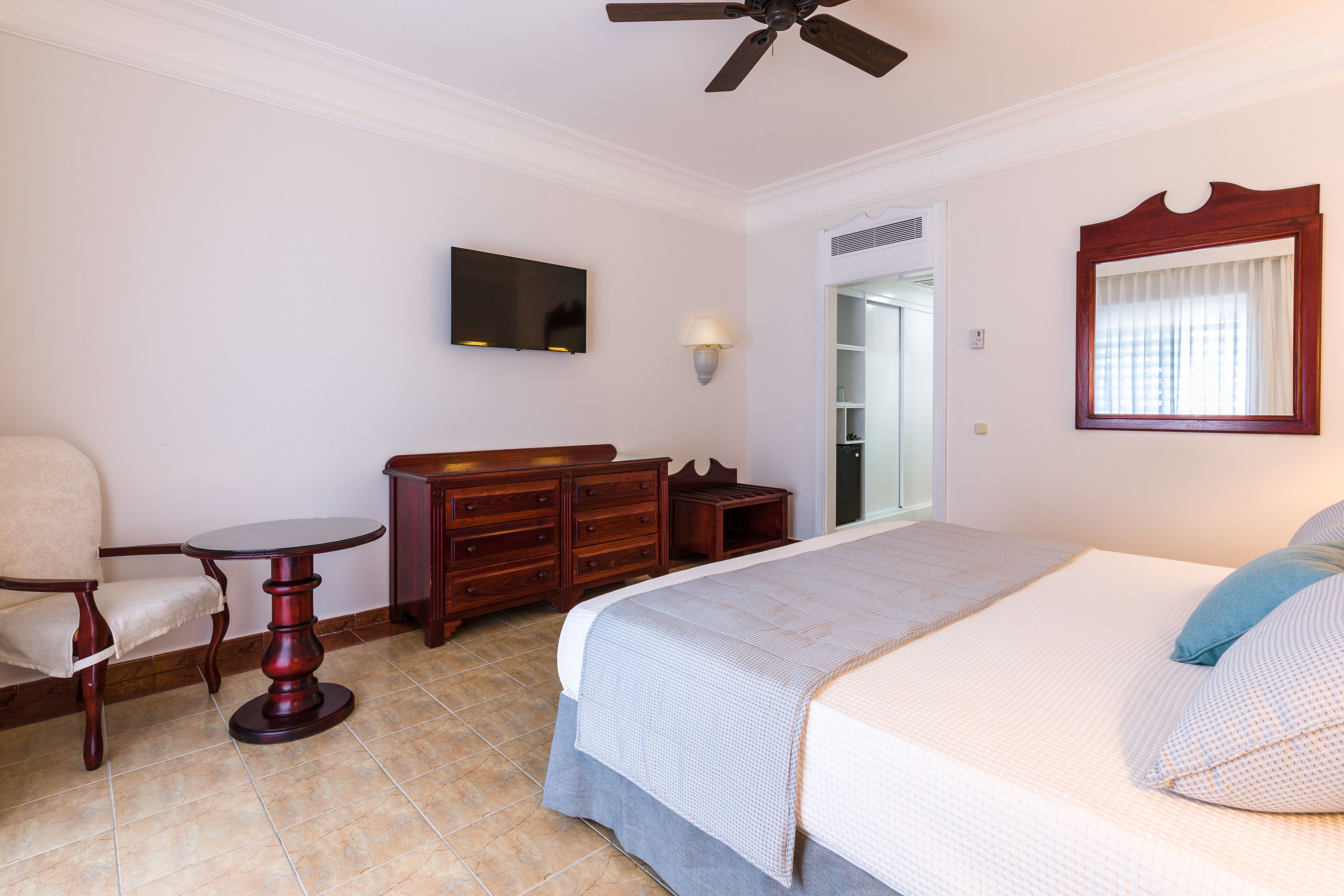 A hotel room with a bed, a chair, a small table, a dresser, a television, and a ceiling fan.