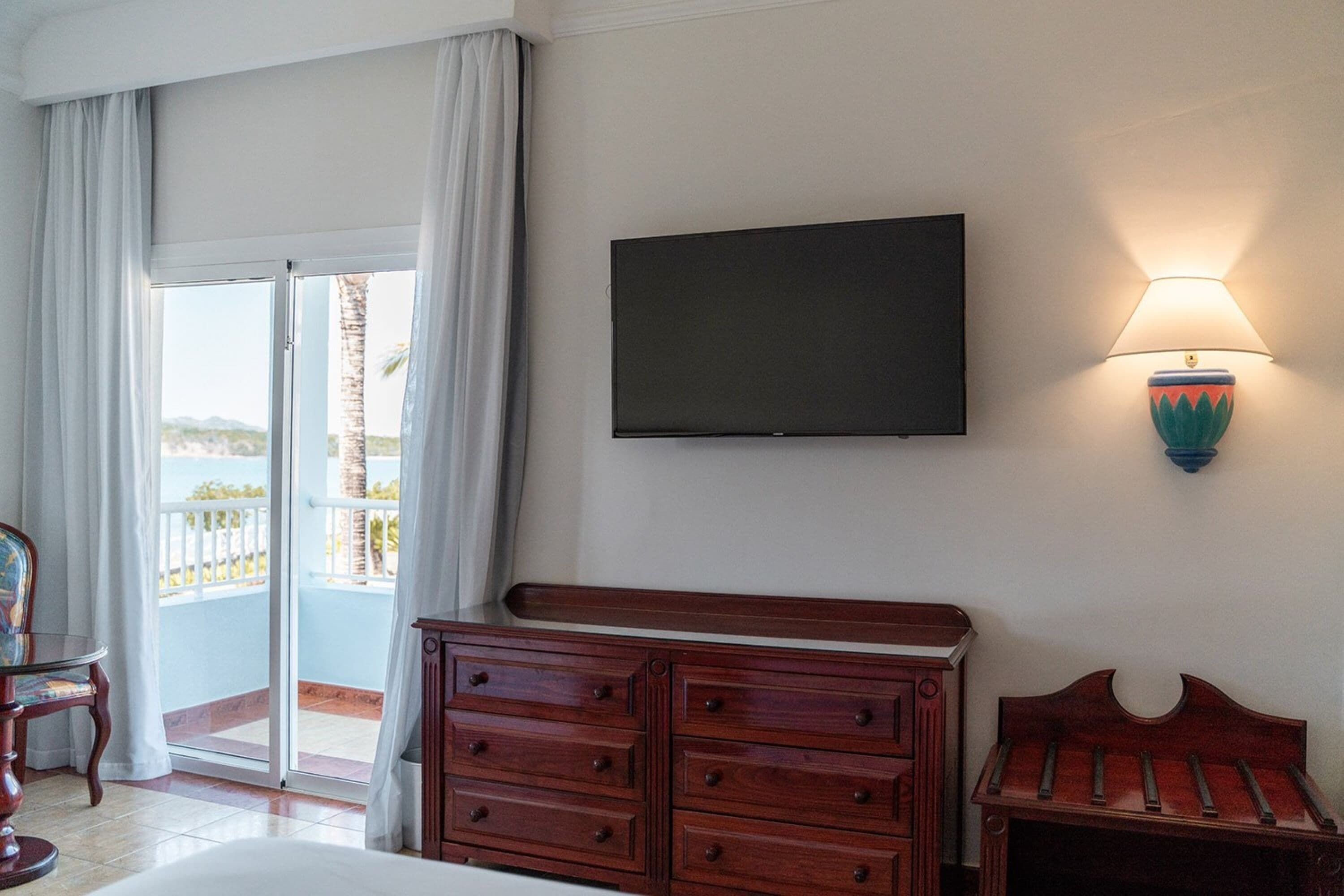 A hotel room with a bed, a wooden dresser, a television mounted on the wall, and a balcony with a view of the sea.