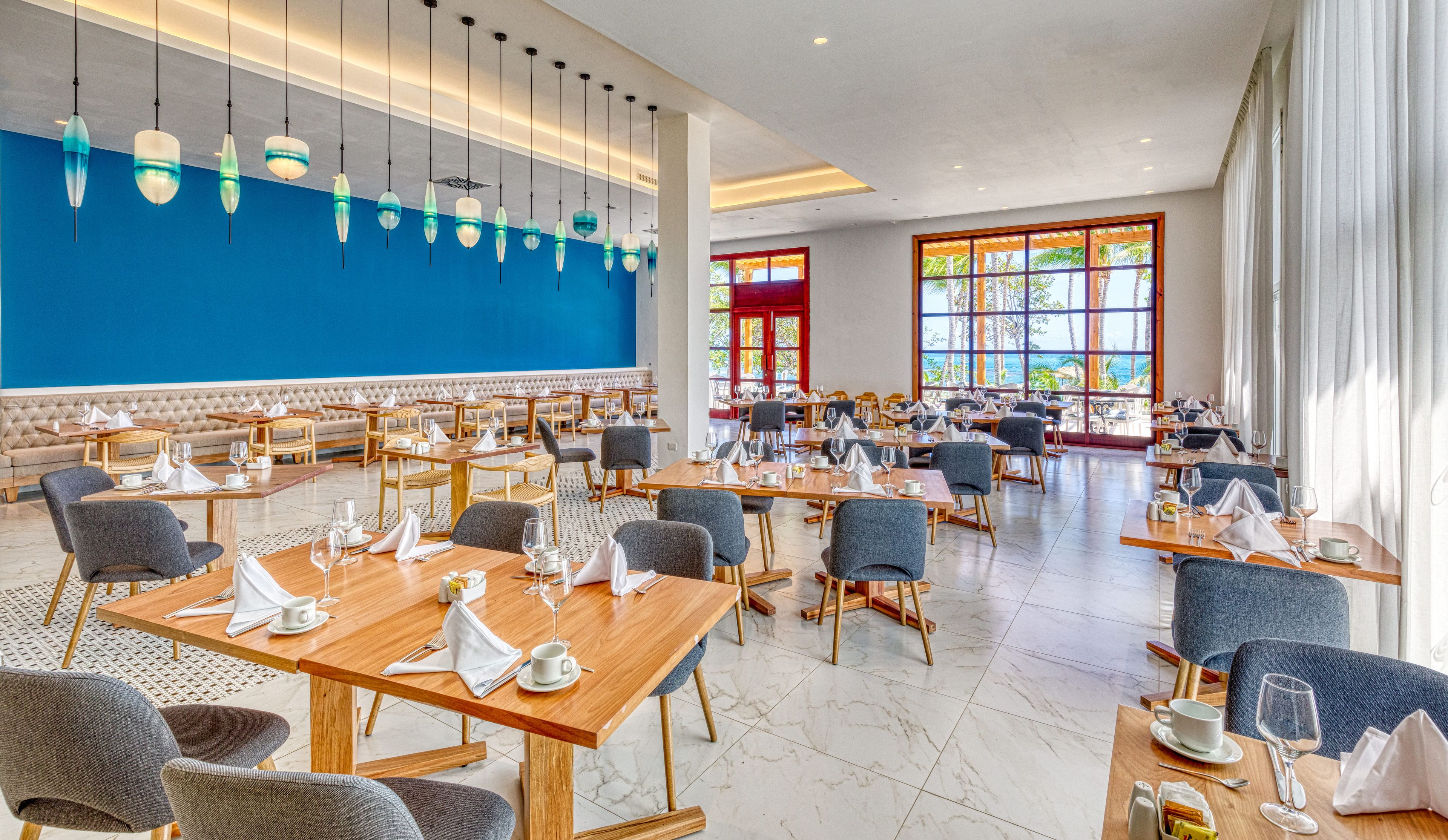 Un comedor espacioso con varias mesas y sillas, un muro azul decorativo y ventanas grandes con vistas al exterior.