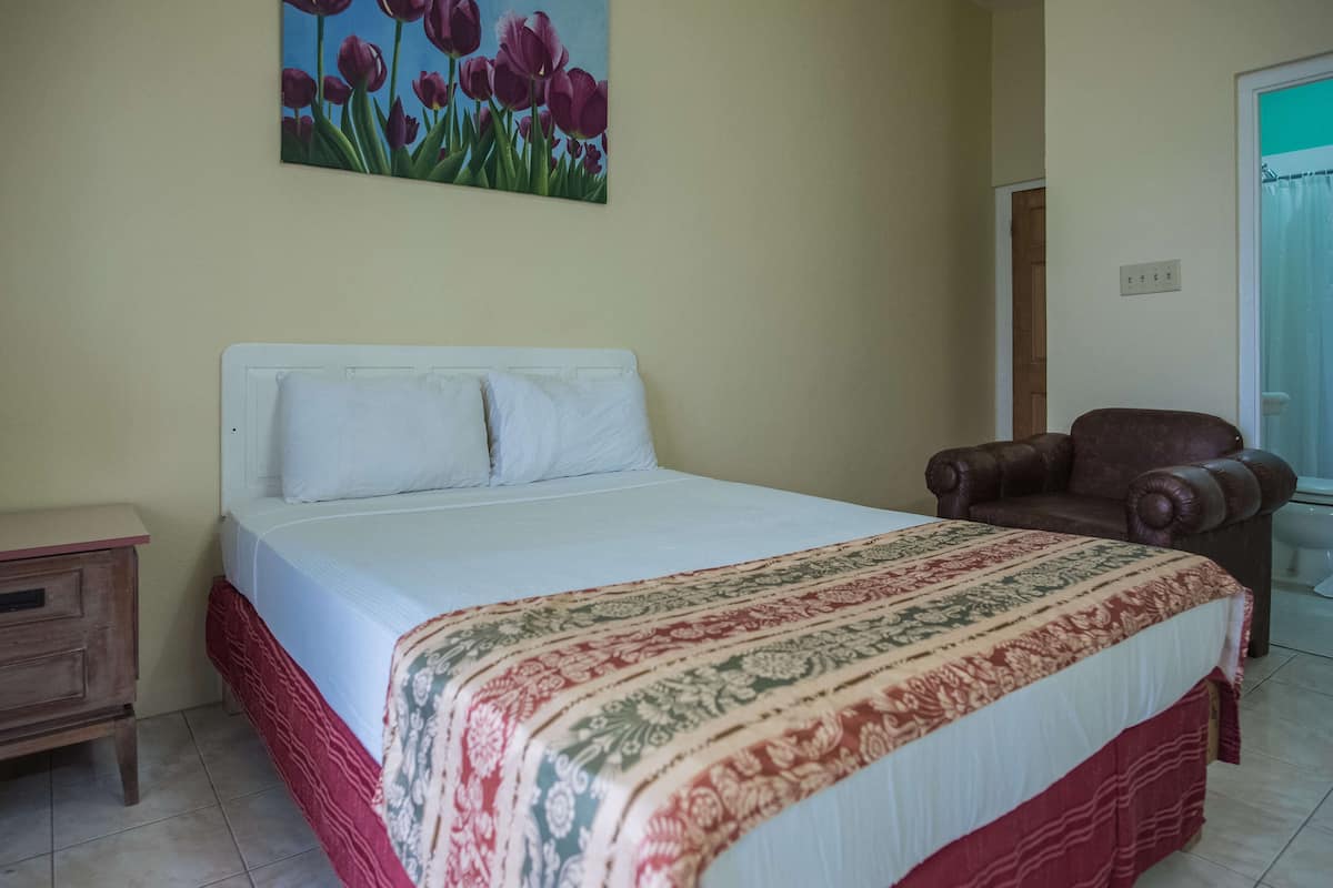 Ein ordentlich bezogenes Bett mit einem gemusterten Überwurf, ein Nachttisch aus Holz mit einer Schublade, ein brauner Ledersessel und ein Gemälde mit Tulpen an der Wand.
