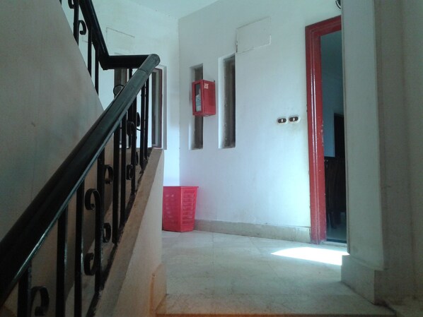 Un escalier avec une rampe métallique mène à un palier. Sur le palier, on trouve une caisse en plastique rouge, et une porte rouge se trouve au fond du couloir.