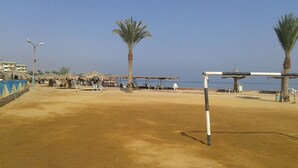 Une plage de sable bordée de palmiers, avec un filet de volley-ball et des constructions aux toits de chaume.
